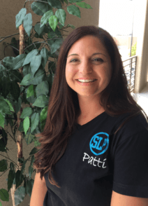 Woman smiling in a black logo T-shirt standing beside leafy plants indoors