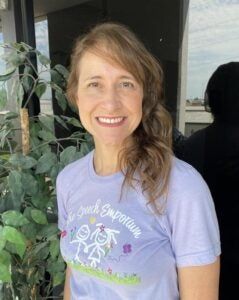 Smiling person in a lavender T-shirt standing indoors beside a plant and window.