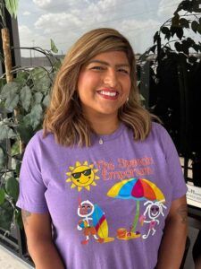 Smiling person in a purple T-shirt with a colorful surf-themed graphic, standing outdoors.