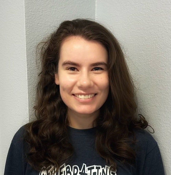 Smiling person with long dark hair in a navy T-shirt against a light gray wall