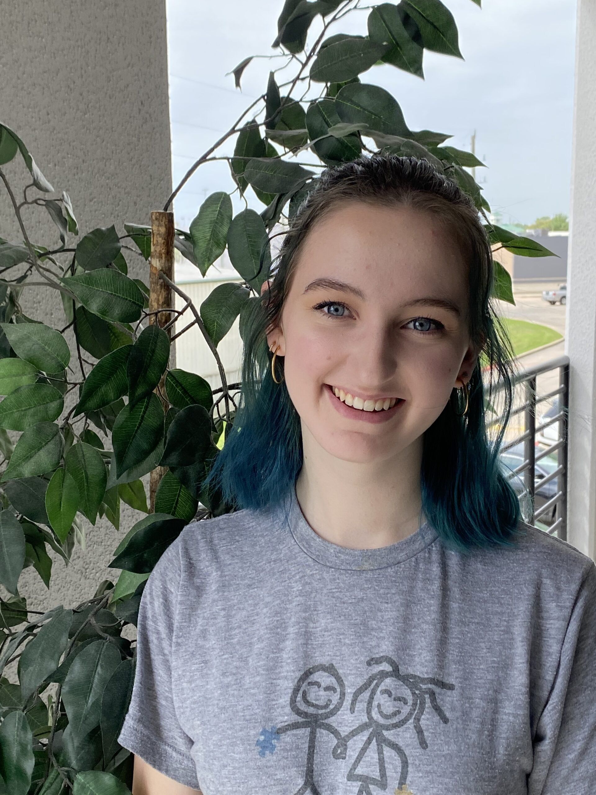 Smiling person with blue-tipped hair standing by a leafy balcony railing in a gray T-shirt