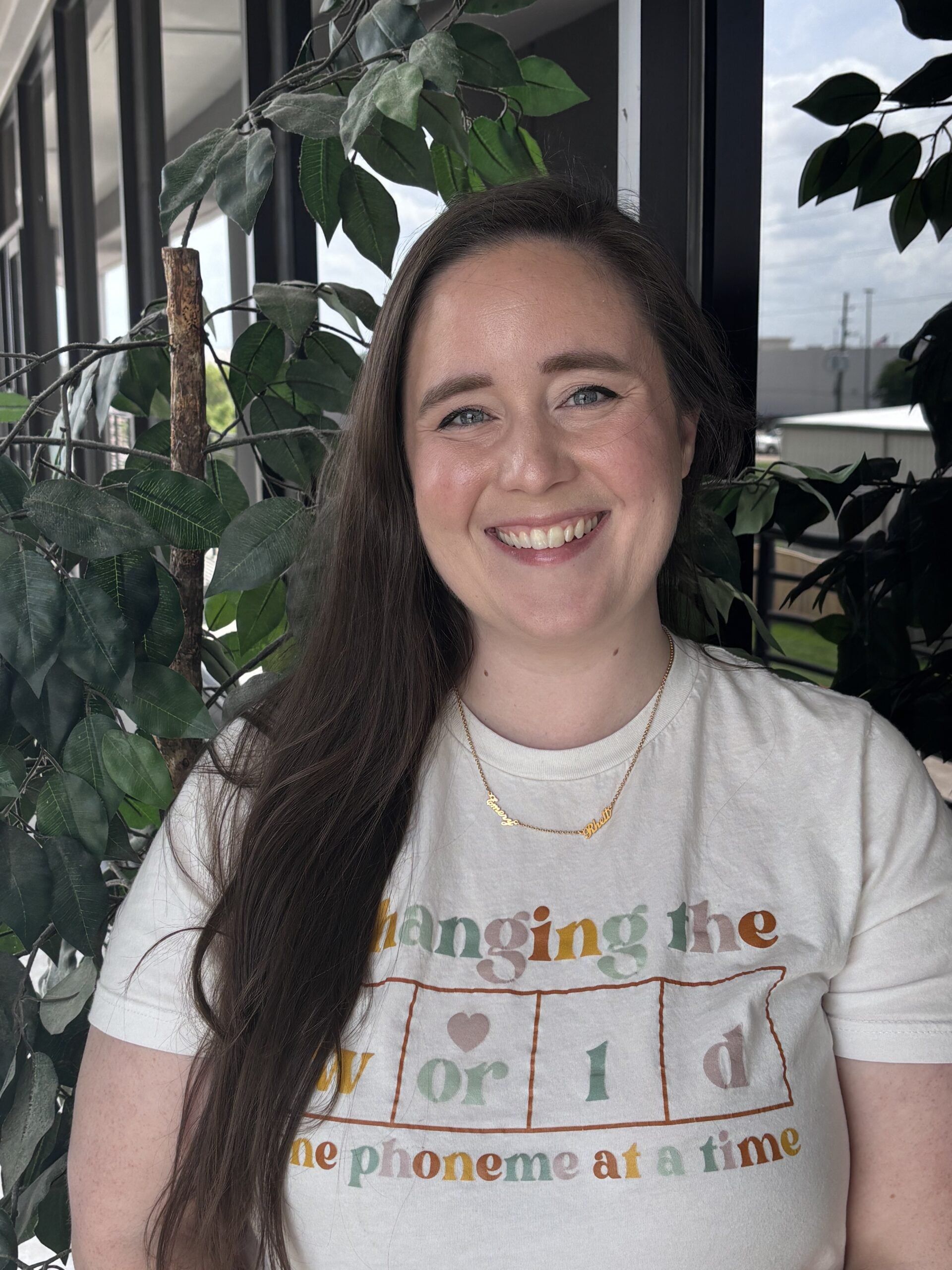 Smiling woman standing indoors beside leafy plants, wearing a white graphic T-shirt.