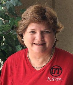 Smiling person in a red shirt with the name Karen, standing indoors near a plant