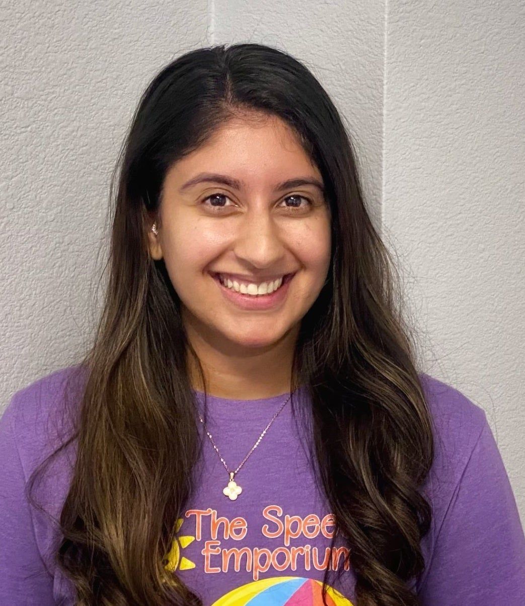 Smiling person in a purple T-shirt standing against a light gray wall.