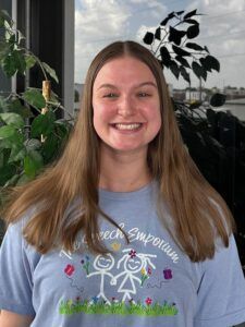 Smiling person in a light blue graphic T-shirt standing outdoors by green plants and a window.