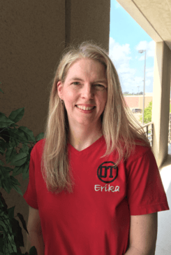 Woman in a red shirt standing indoors and smiling near a window.