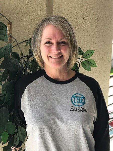 Smiling person in a gray and black shirt standing indoors beside a leafy plant