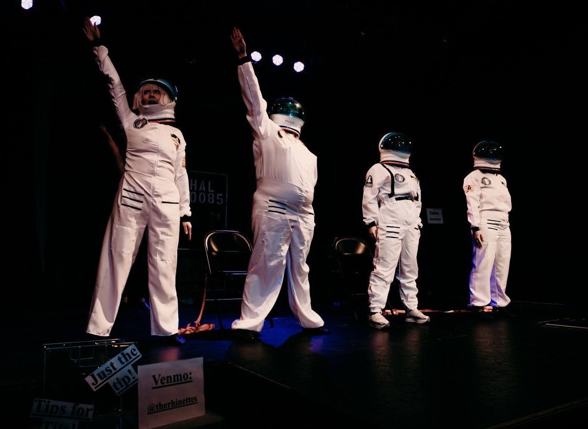 Four people in white astronaut suits on a dark stage with arms raised.