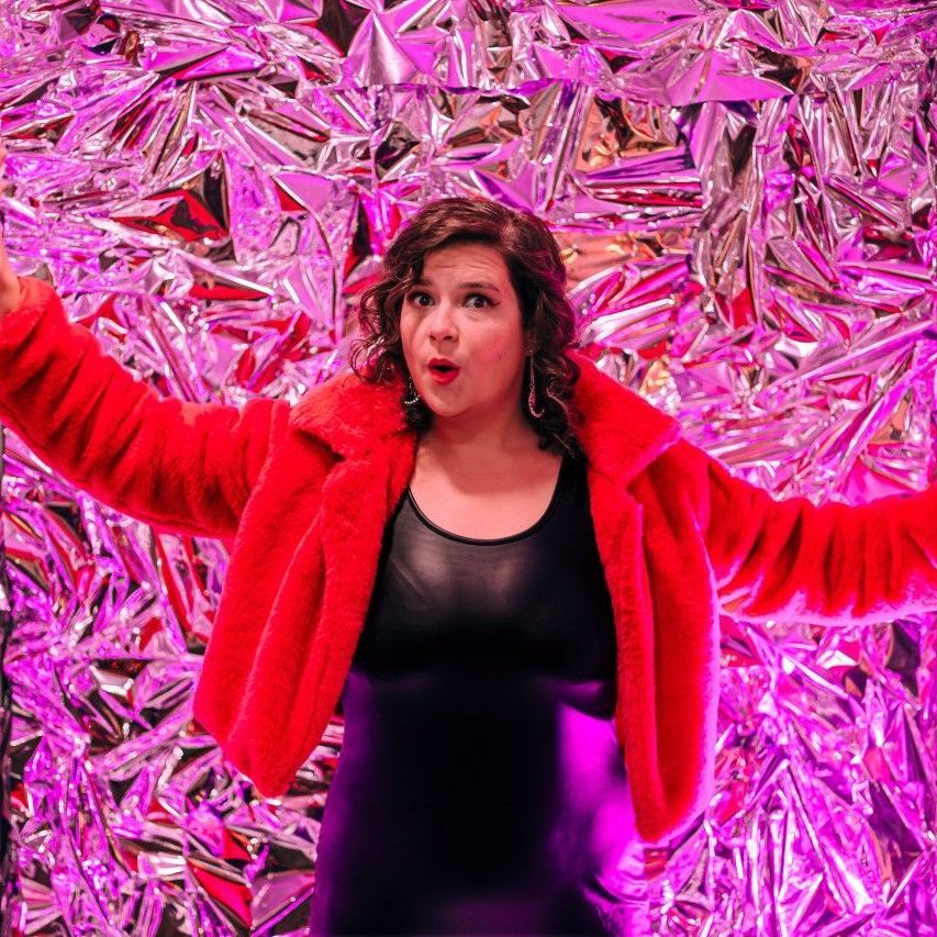 A woman in a red fur coat is standing in front of a pink and purple background.