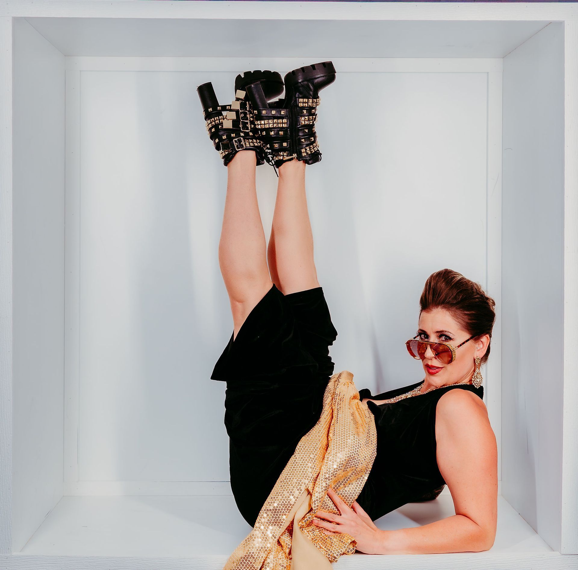 Woman in black outfit, gold scarf, sunglasses, and heeled boots, upside down in a white box.
