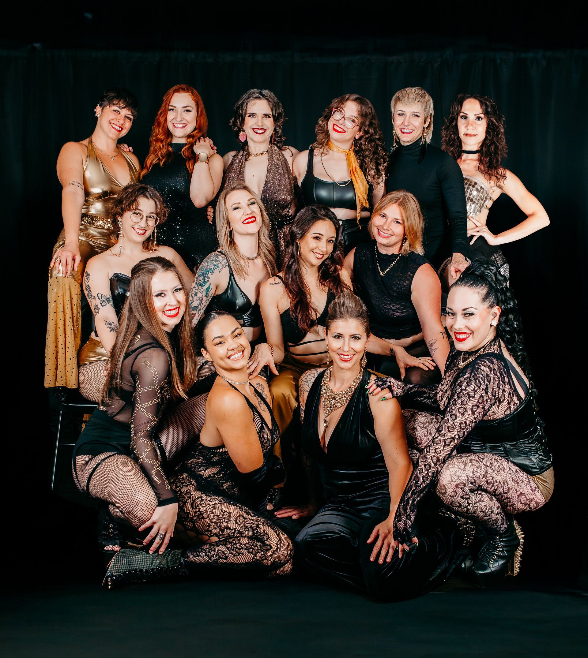 Group of women posing together, wearing diverse outfits, smiling, against a black backdrop.