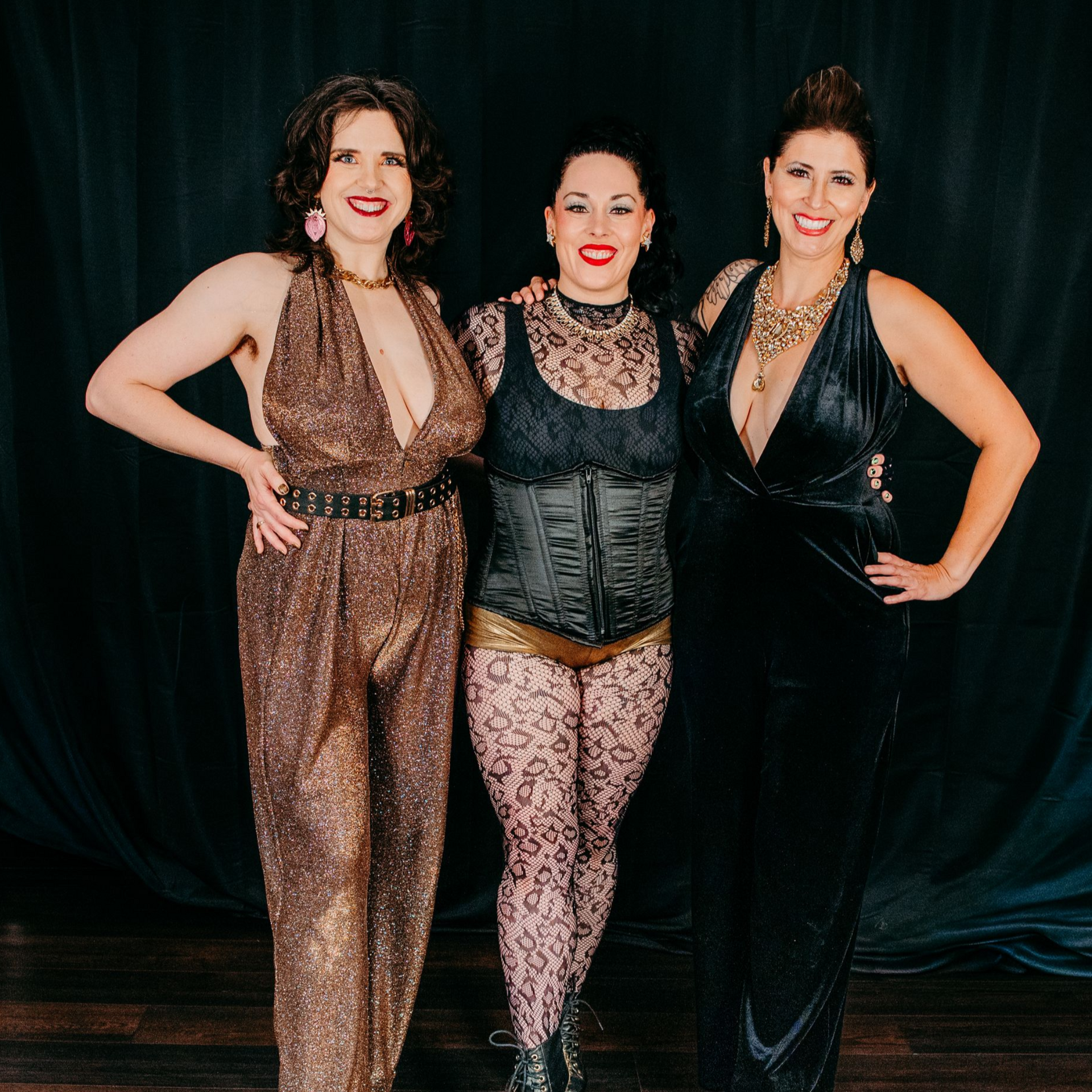 Three women in glamorous outfits posing in front of a black curtain.