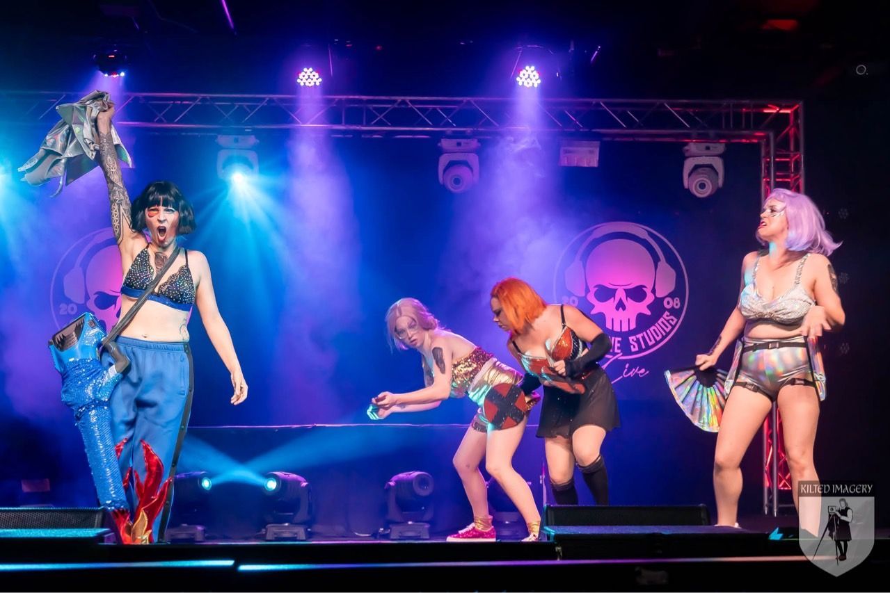 A group of women are dancing on a stage with a skull in the background.