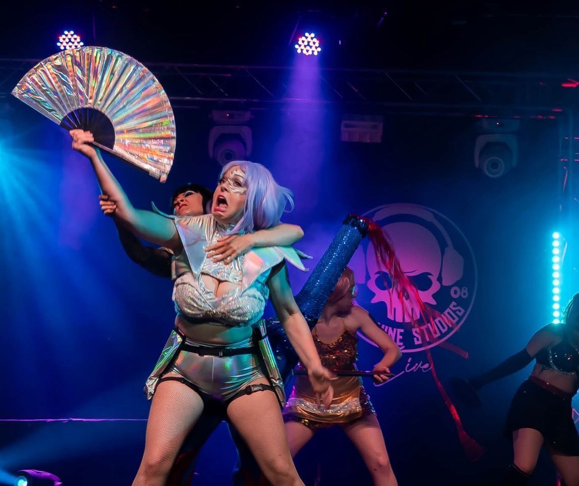 A performer on stage with a fan, being embraced by another. Stage is lit with blue and purple lights.