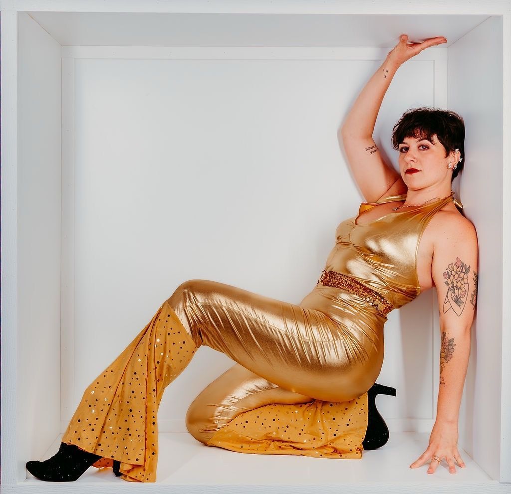 Woman in gold jumpsuit, posed in a white box, one arm raised.