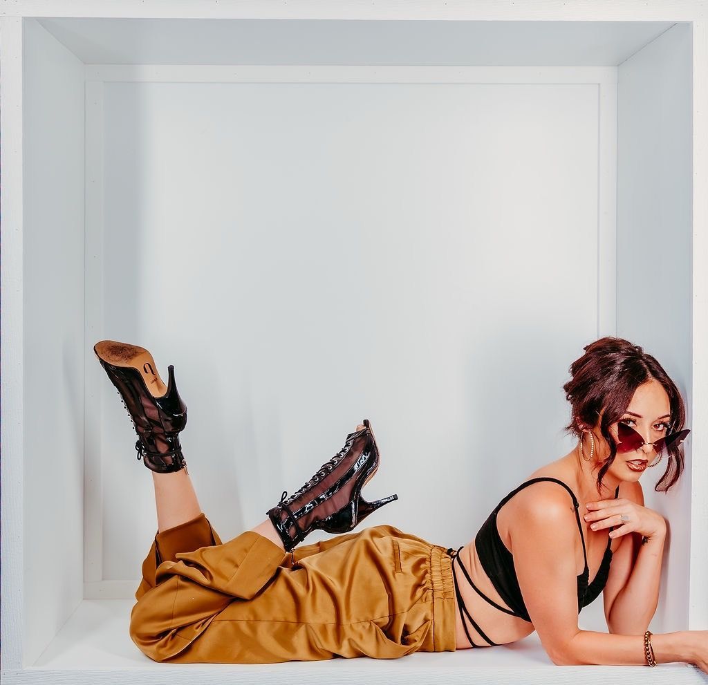 Woman in brown pants and black top reclines in a white box, wearing high-heeled boots and sunglasses.