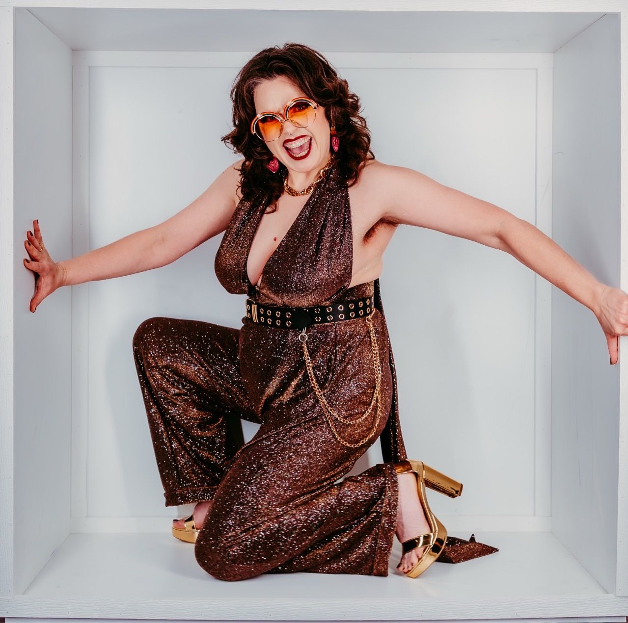 Woman in sparkly brown jumpsuit kneels in a white box, arms outstretched, smiling, wearing glasses and gold heels.