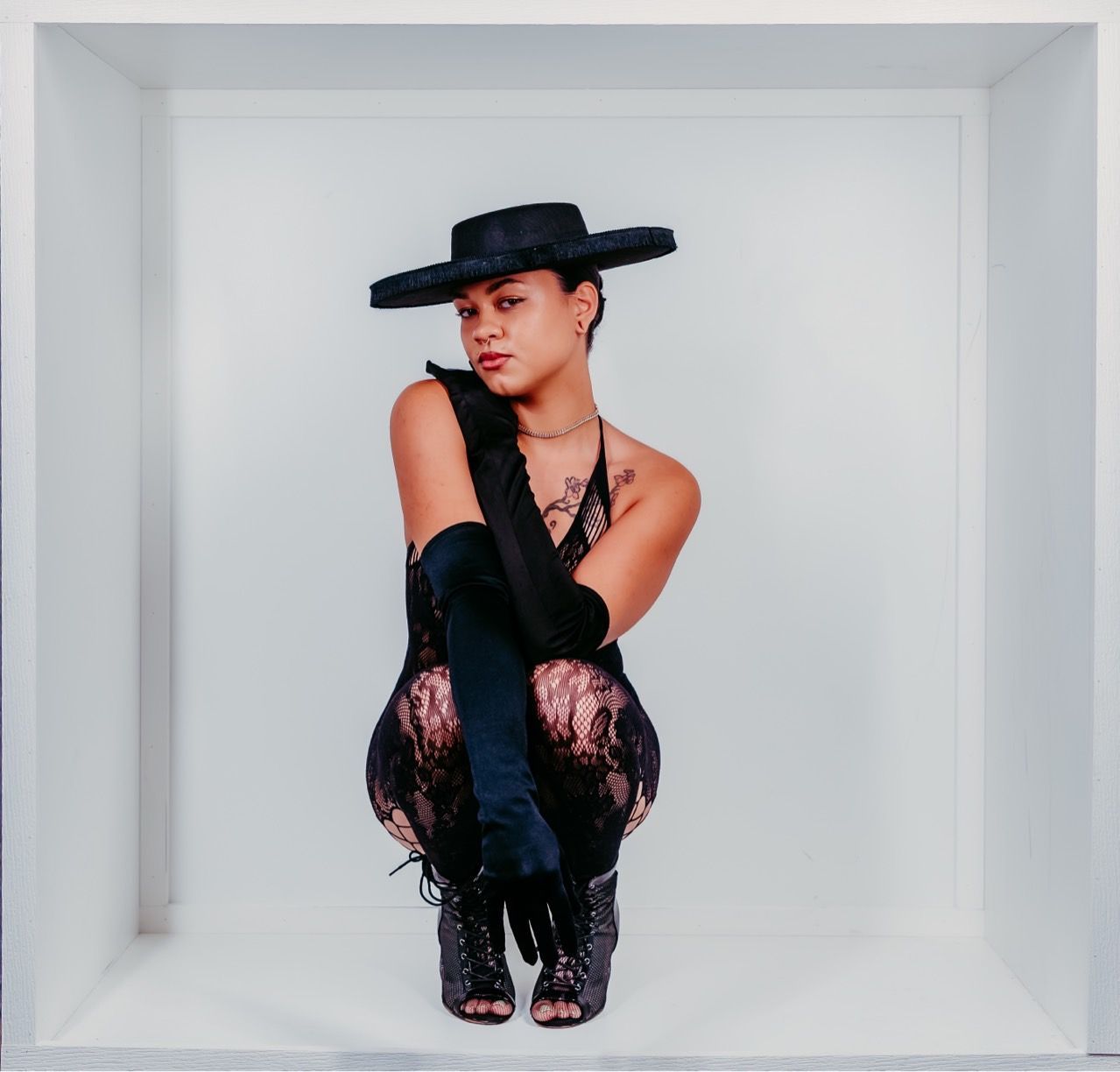 Person in black outfit, wide-brimmed hat, gloves, and boots squats in a white box, gazing at the viewer.
