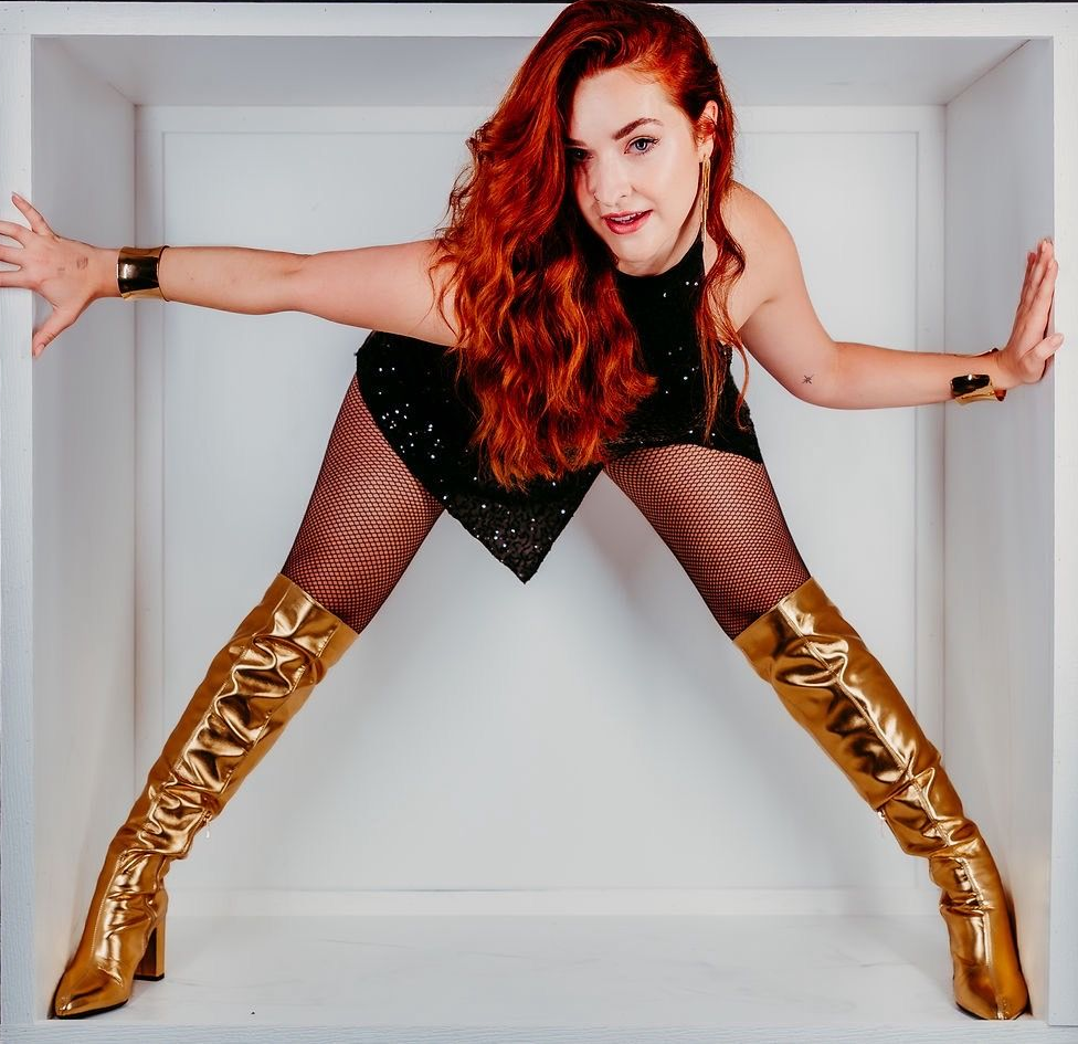 Woman  in a small box, arms and legs out, wearing a black dress, fishnets, and gold boots.