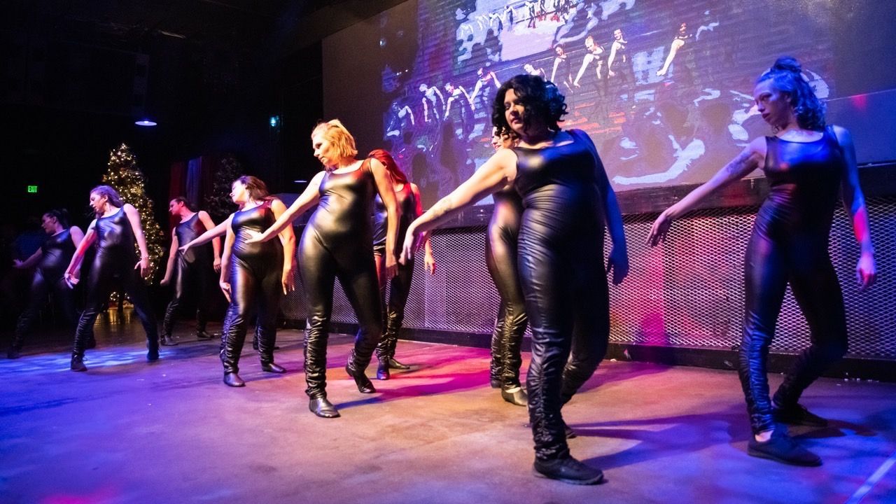 Dancers in black outfits perform on a stage with a projected backdrop. Blue and purple stage lights.
