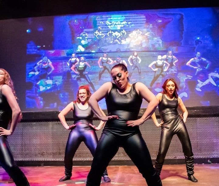 Dancers in black outfits on stage, posing. A group of dancers is visible on a screen behind them.