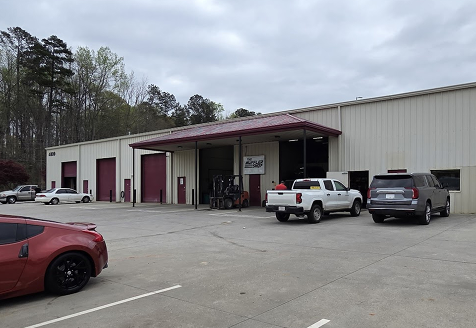 Warehouse loading dock with several parked trucks and cars outside a large industrial building. | The Muffler Shop