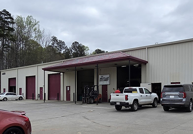 Industrial warehouse building with open loading bay, parked trucks, and overcast sky | The Muffler Shop