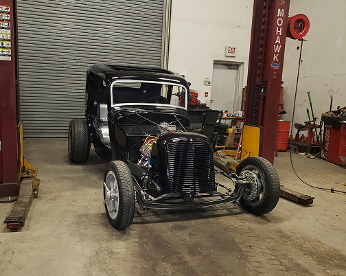 Black hot rod in an auto shop, front view with exposed wheels and engine, near a closed garage door. | The Muffler Shop