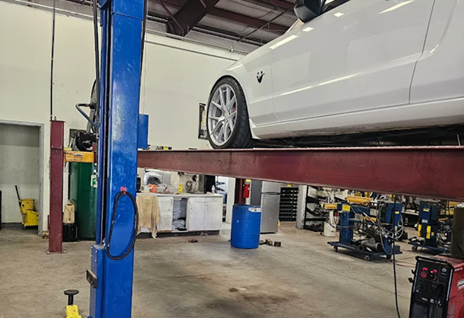 White car on a lift in an auto repair shop with tools and equipment below. | The Muffler Shop