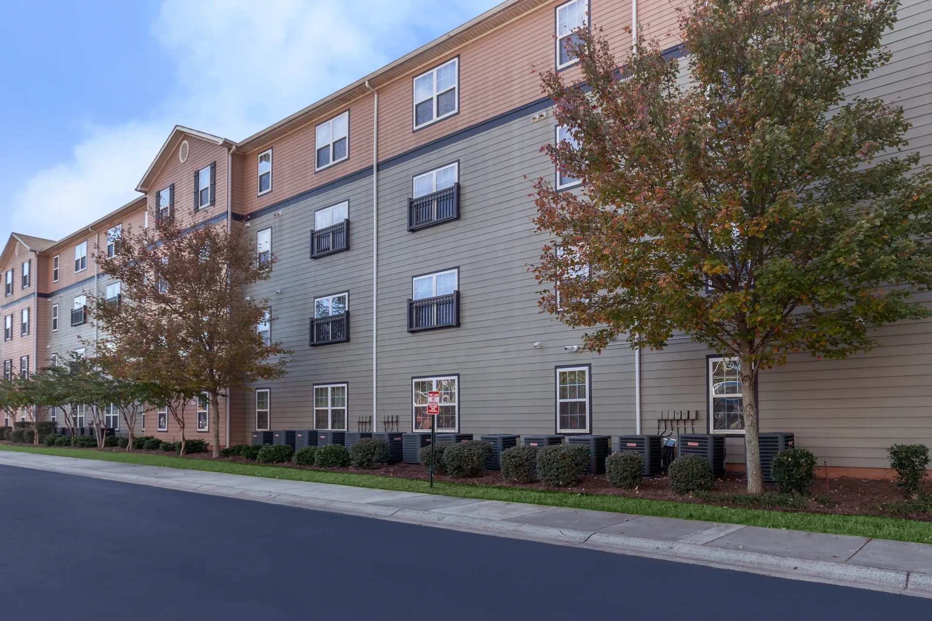 Exterior view of a multi-story apartment building with green and tan siding, surrounded by trees in autumn colors.