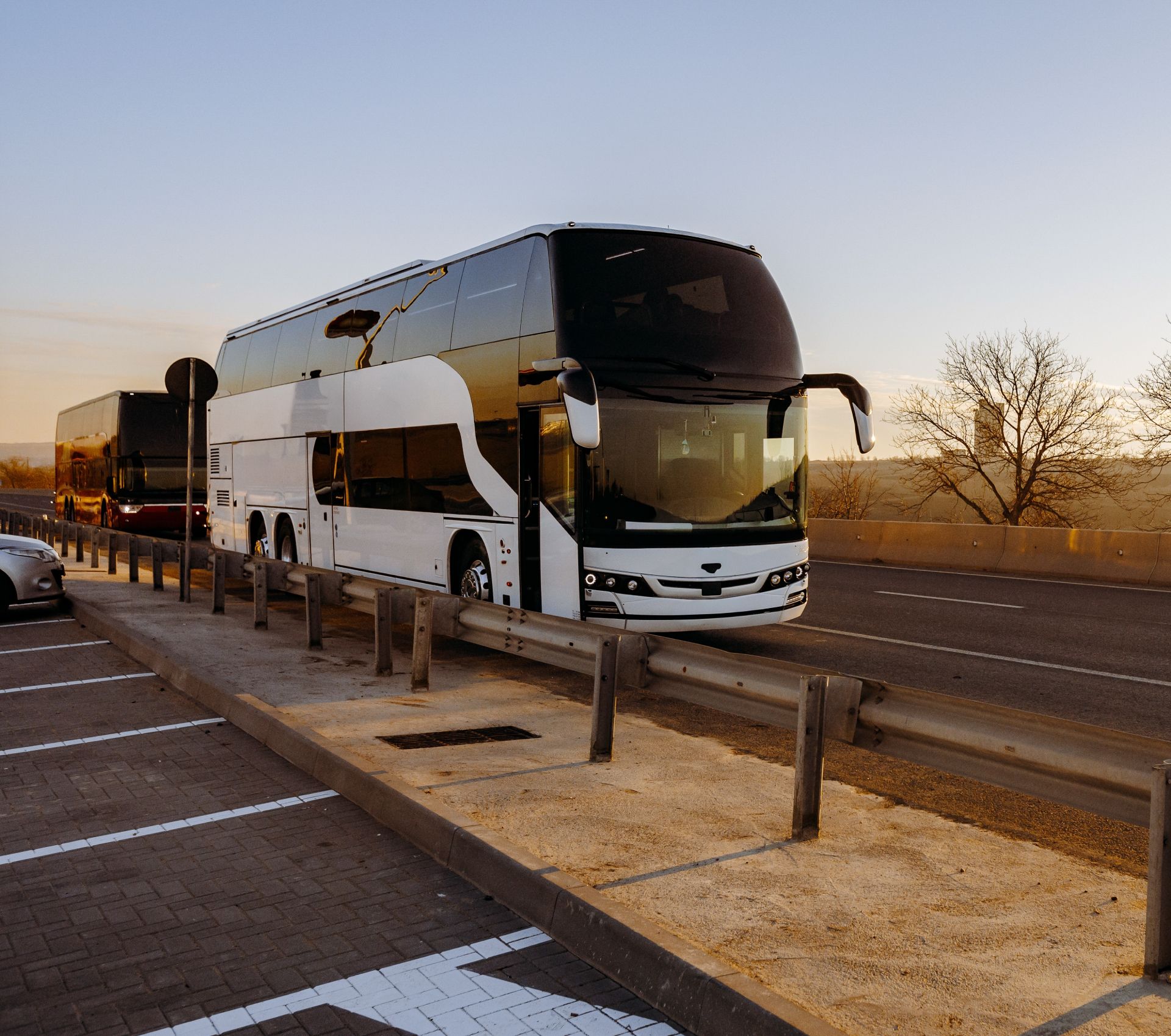 Two-tone coach bus parked on a roadside pull-off at sunset, with desert trees and a clear sky.