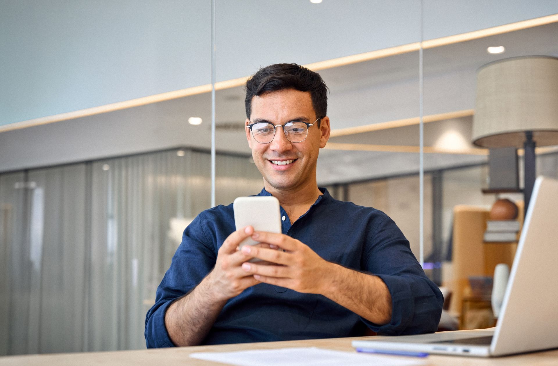 Smiling person in a blue shirt using a smartphone at a desk with a laptop in a bright office