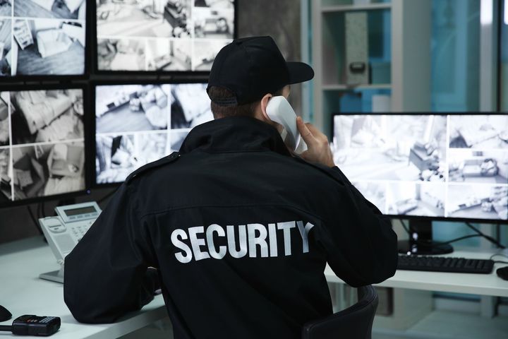 Security guard in uniform at a desk, monitors displaying surveillance footage. He's talking on a phone.