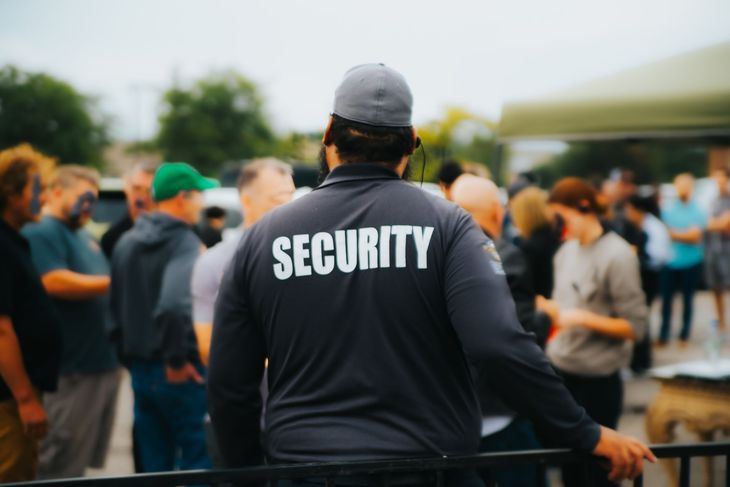 Security guard in gray shirt, standing in front of a crowd of people.