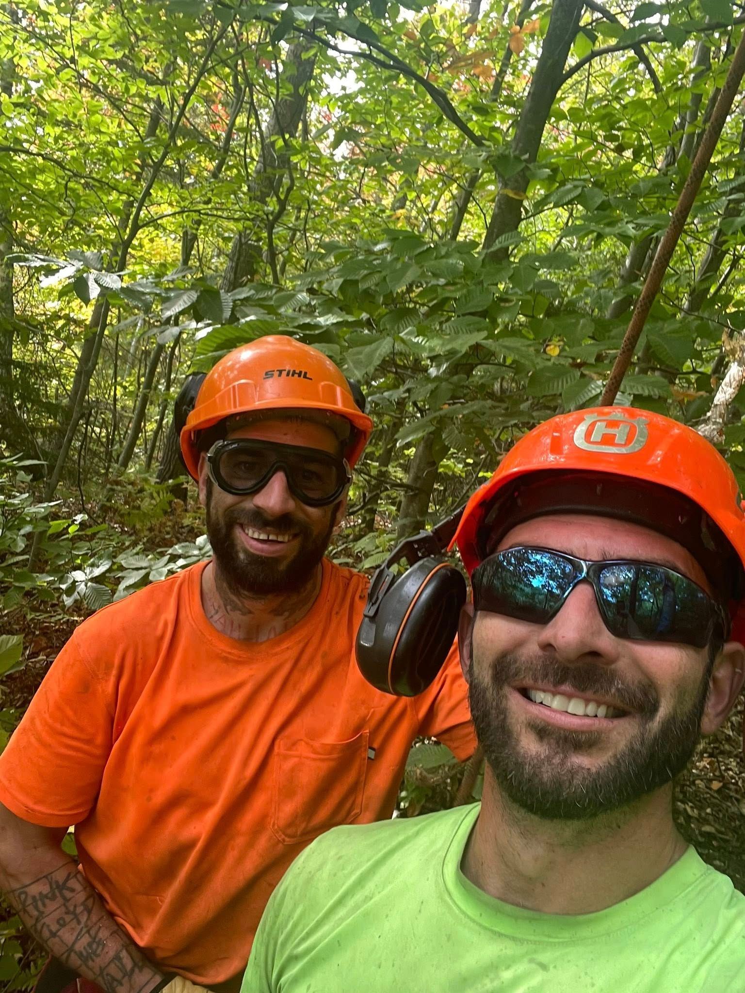 Deux ouvriers souriants, portant des casques orange et des lunettes de sécurité, se tiennent au milieu d'une forêt verdoyante.