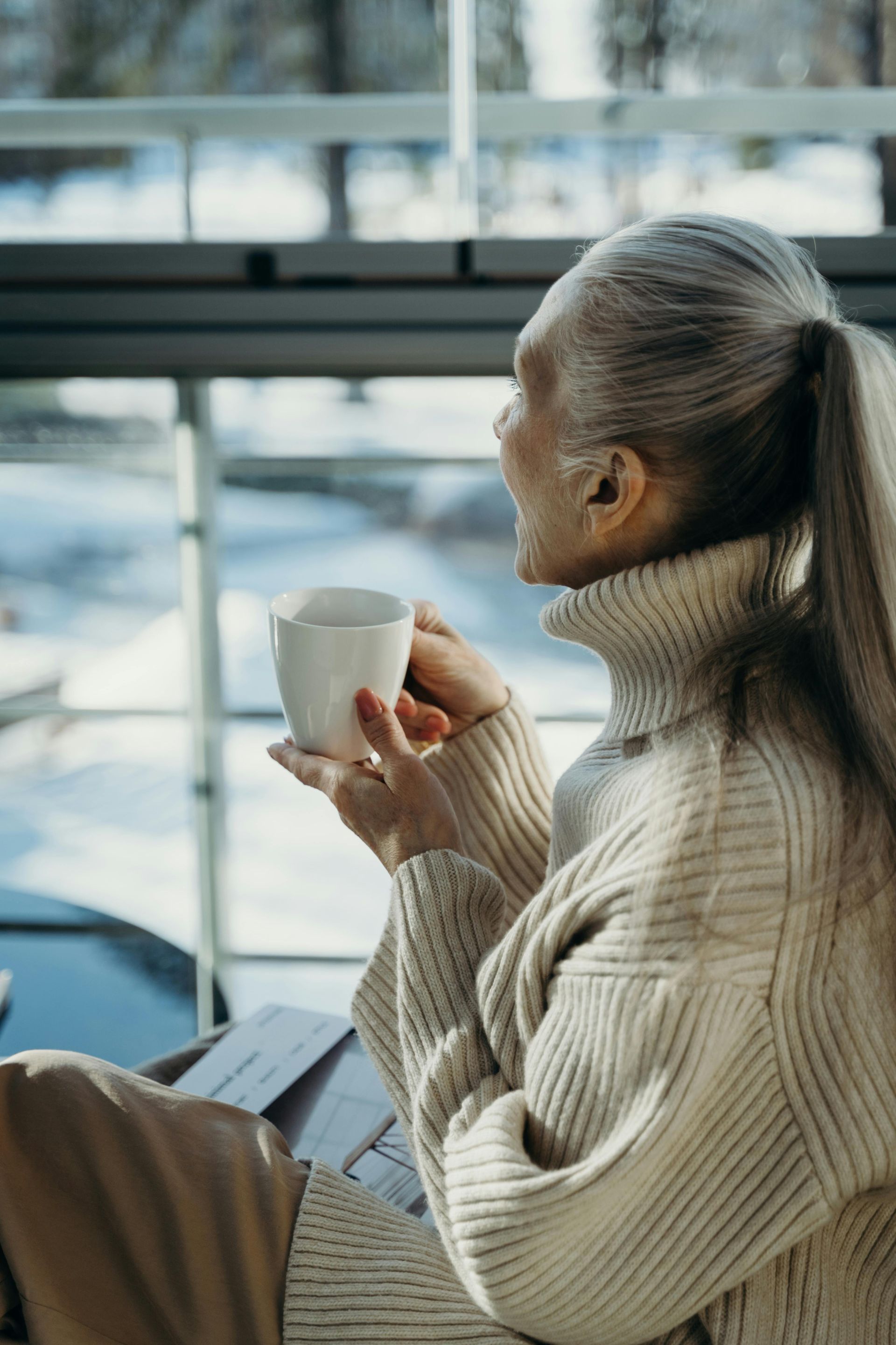 En kvinde i en sweater sidder foran et vindue og holder en kop kaffe.