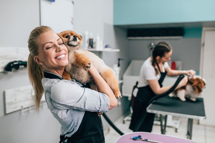 Groomer smiling while holding a small dog in a bright pet grooming salon, with another dog on a table behind.