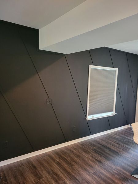 Dark paneled wall with a window, outlet, and diagonal lines. Wooden floor and white ceiling beams.
