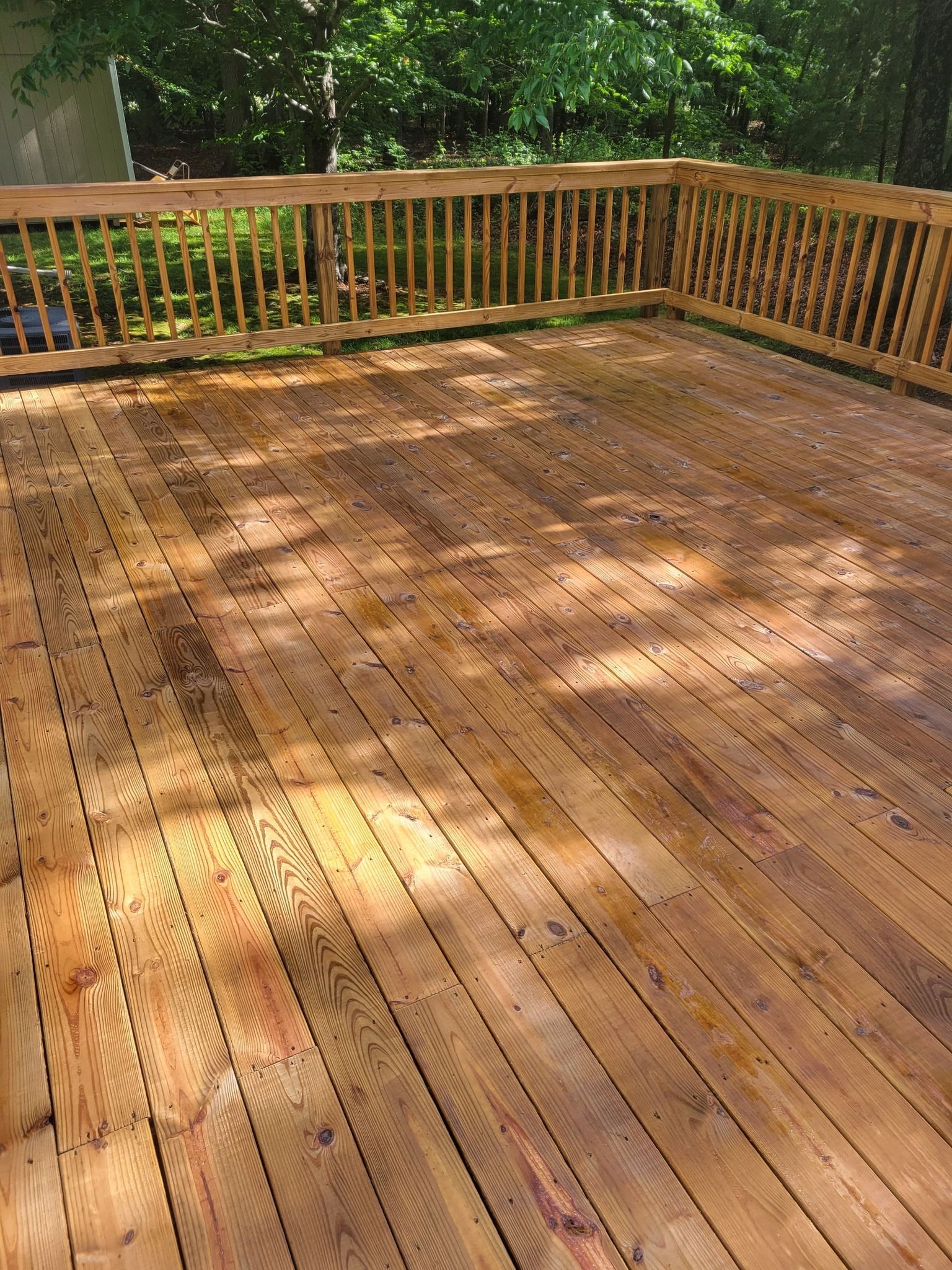 Wooden deck with railings, viewed from above. Sunlight dappled across the boards. Trees in the background.