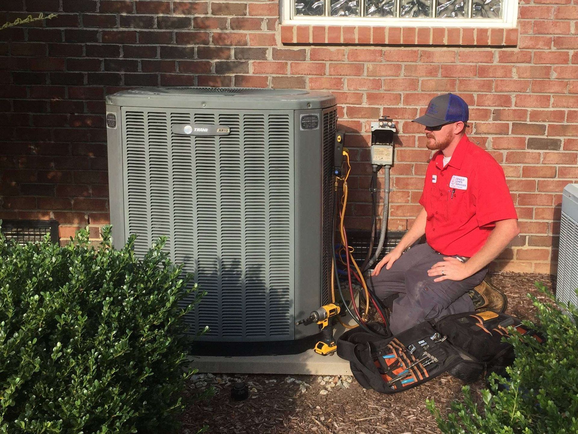 Technician kneels beside an outdoor HVAC unit, inspecting equipment next to a brick house.