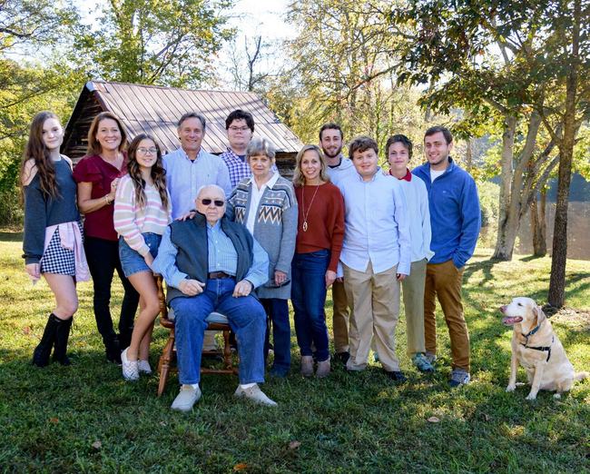 Group of people posing outdoors in a sunny yard with a house, with a dog sitting at the right.