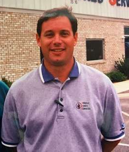 Man in a gray polo shirt smiling outdoors in front of a brick building