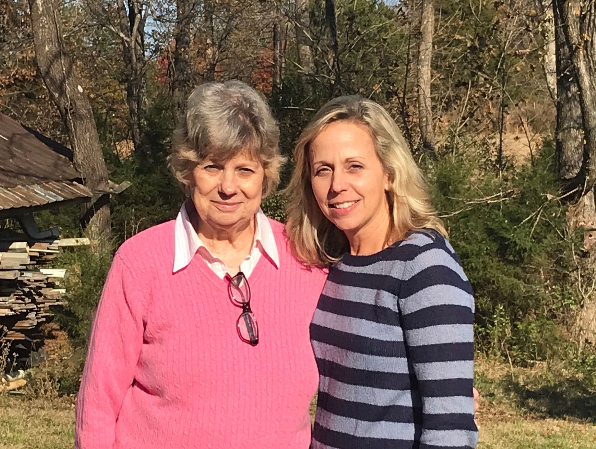 Two women standing outdoors in a wooded area, smiling at the camera.