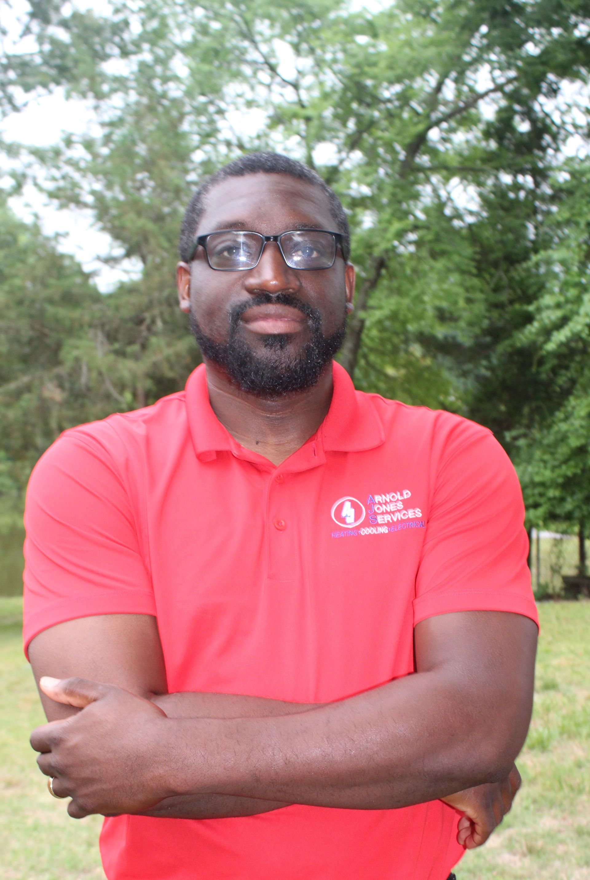 Man in a red polo shirt standing outdoors with arms crossed in front of green trees