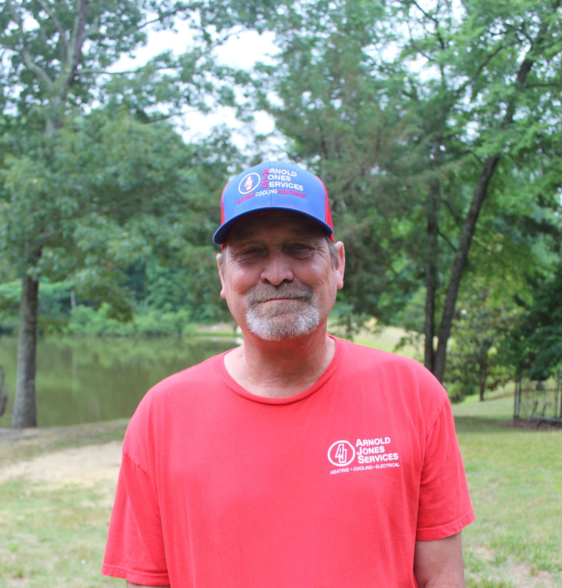 Man in a red shirt and blue cap standing outdoors near a pond and trees