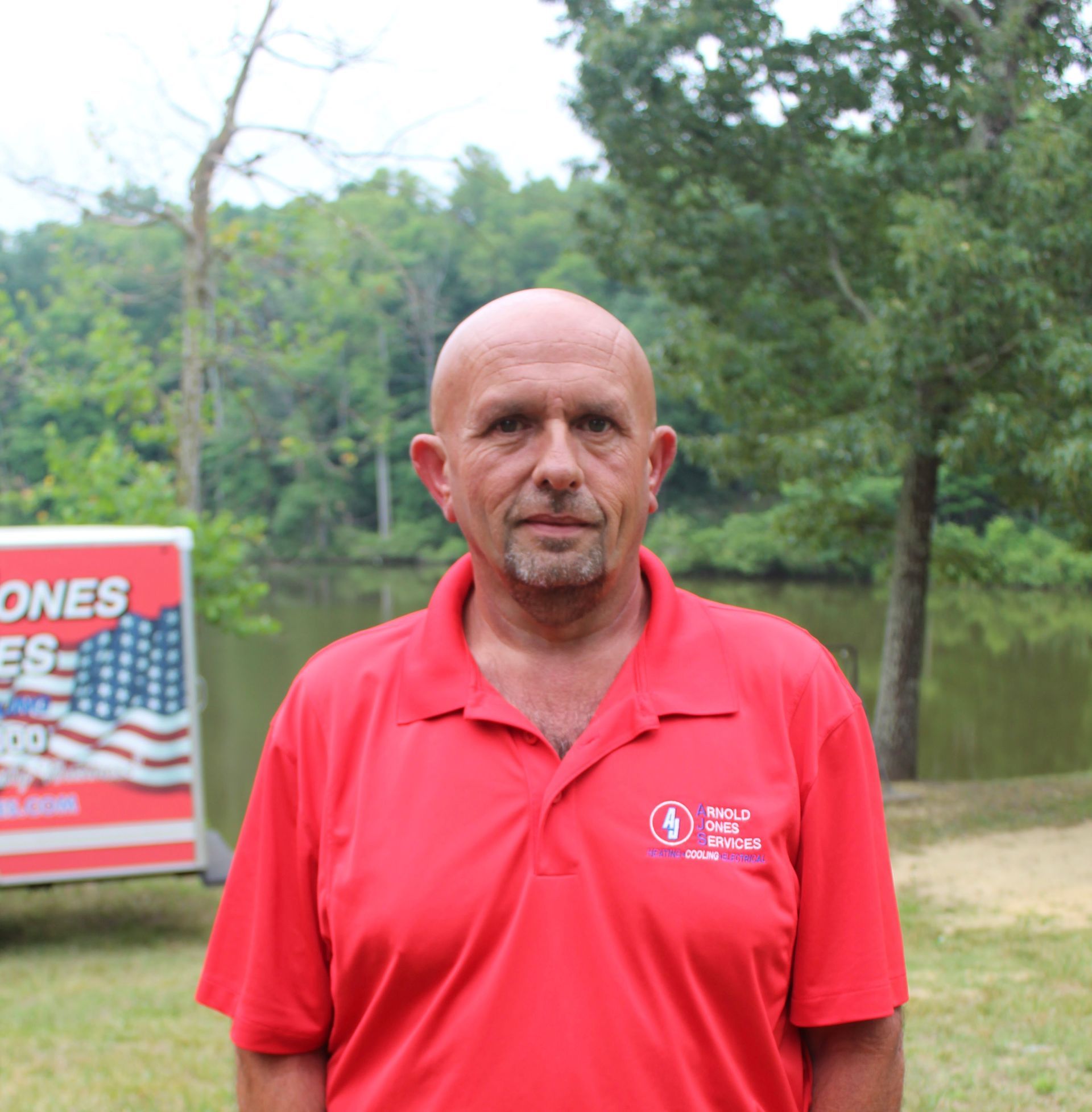 Man standing outdoors in a red polo shirt with trees and a sign in the background