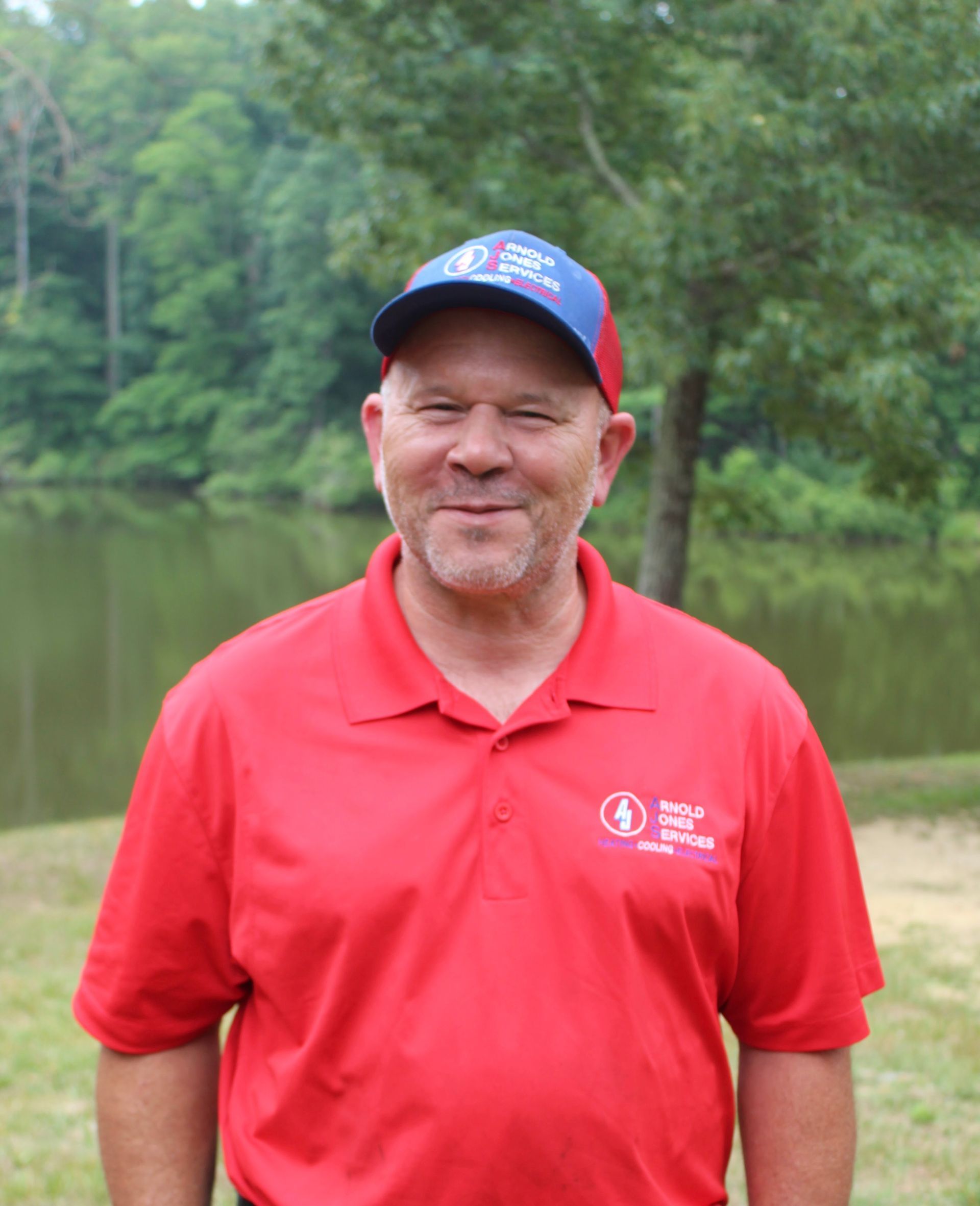 Man in red polo and baseball cap standing outdoors by a lake, smiling at the camera