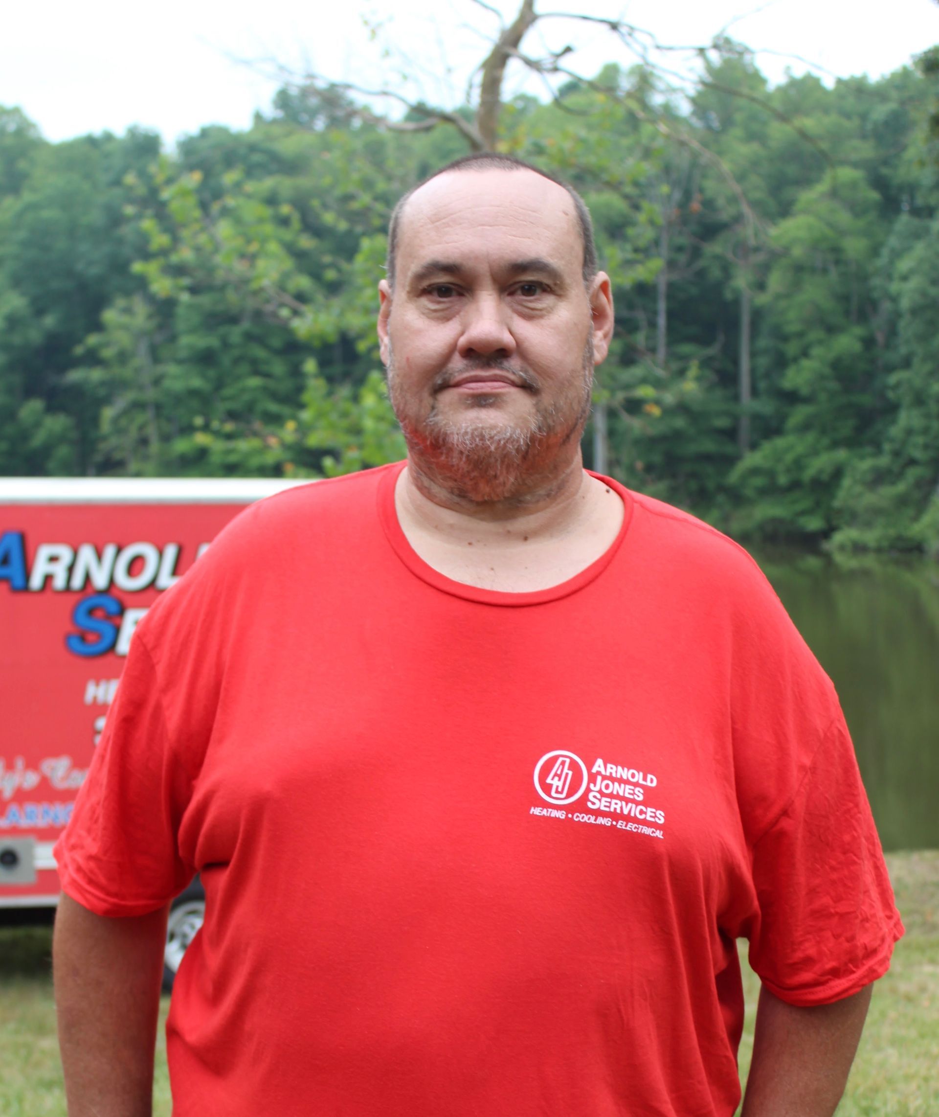 Man in a red T-shirt standing outdoors by a lake with trees in the background