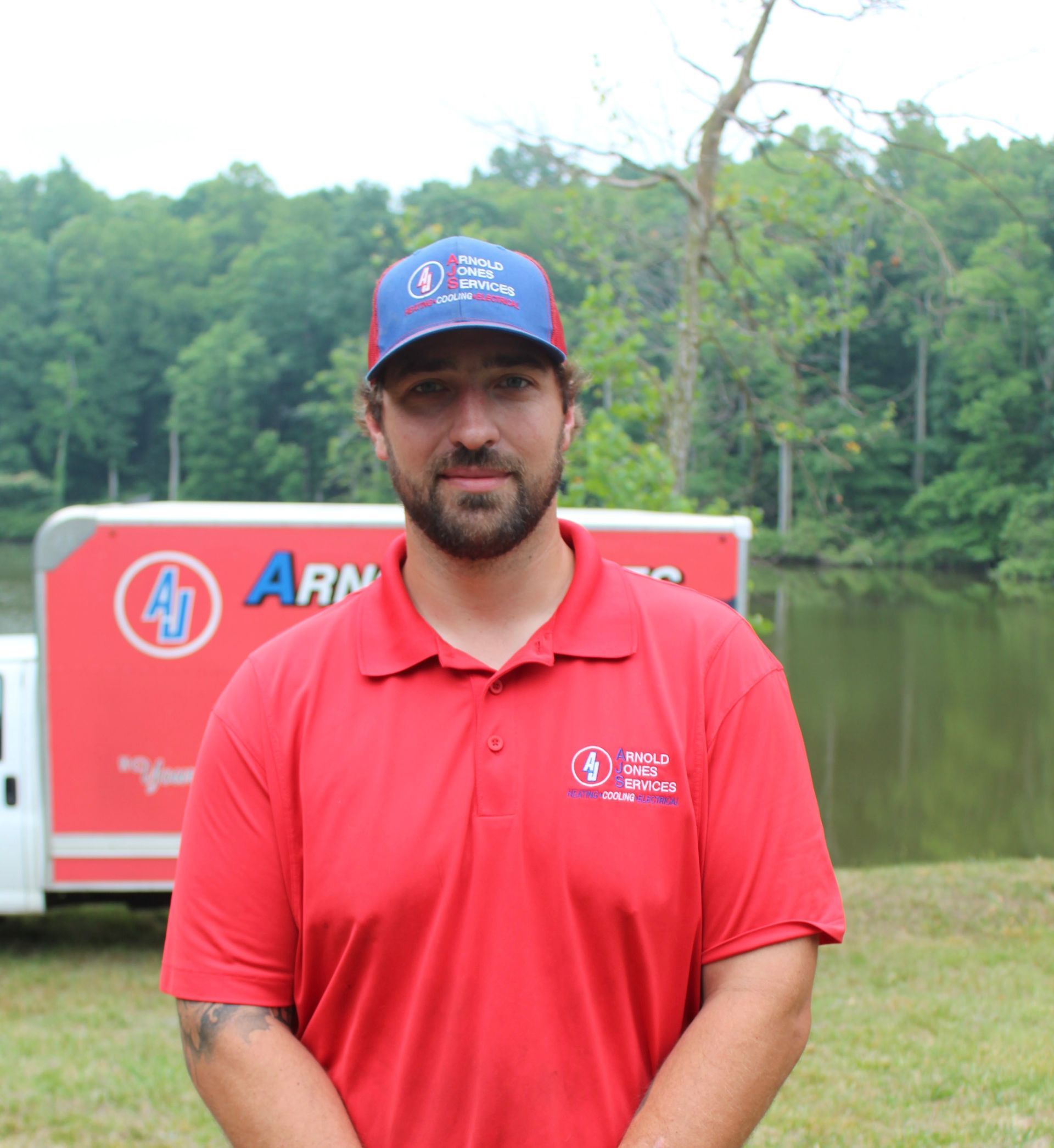 Man in red polo and blue cap standing outdoors by a lake with a red vehicle in the background
