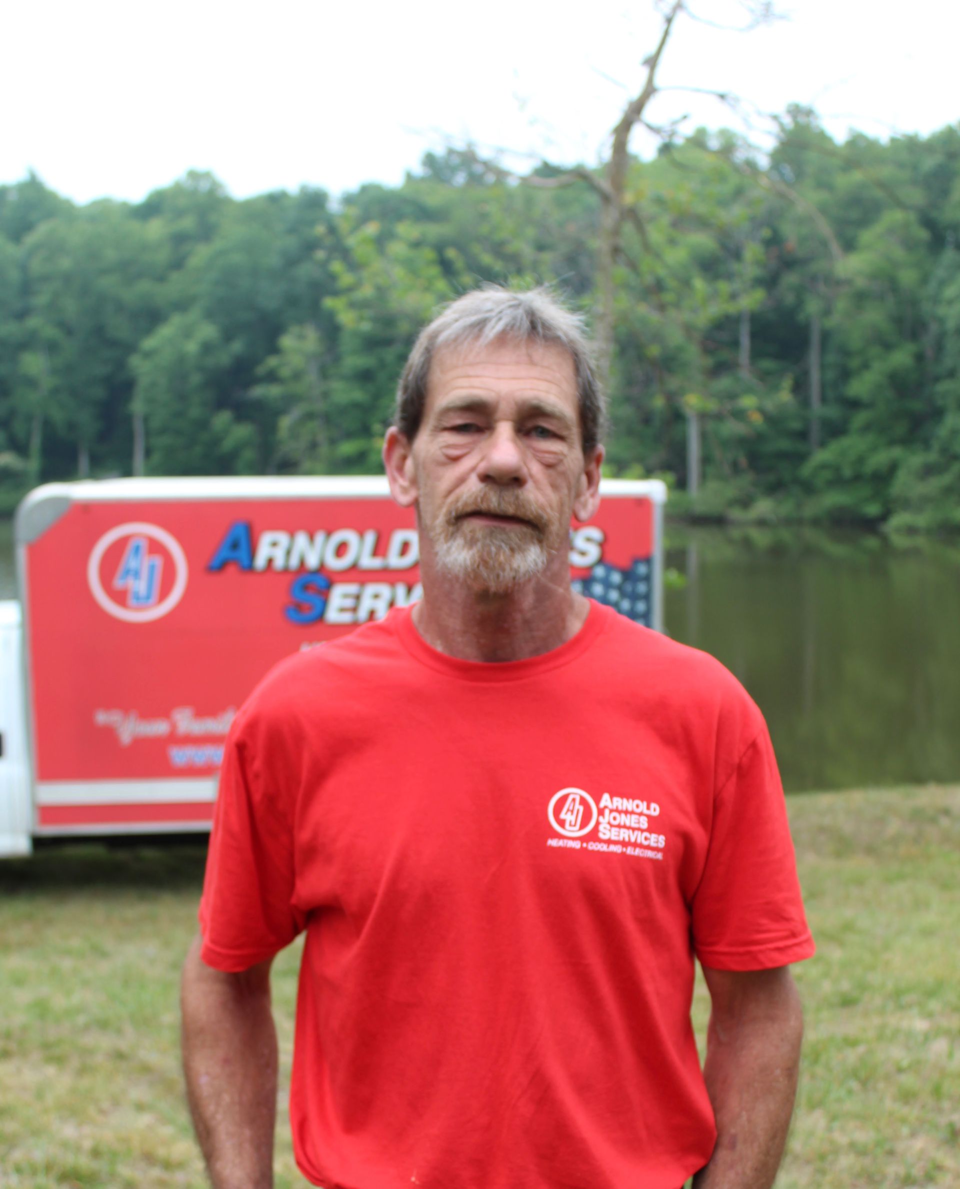 Man in a red T-shirt standing outdoors in front of an Arnold sign and a wooded pond area